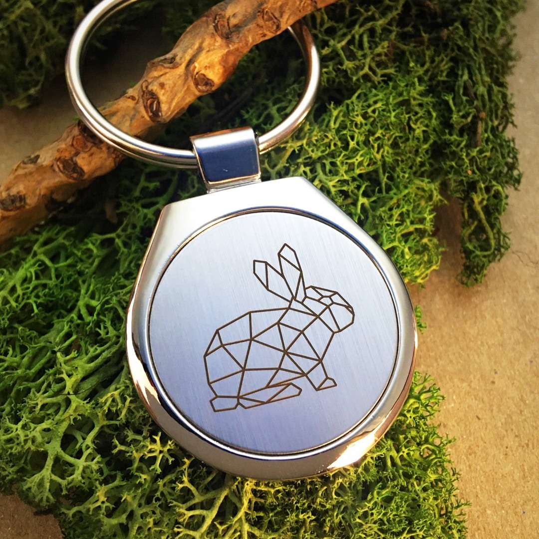 Brelok z metalu z geometrycznym wzorem królika, umieszczony na mchu i gałązce.