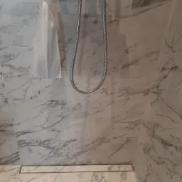 Szczegółowe ujęcie kabiny prysznicowej wyłożonej białym marmurem z szarymi żyłami, widoczny chromowany panel prysznicowy z wężem, mała biała torba zawieszona na panelu oraz odpływ liniowy w podłodze.