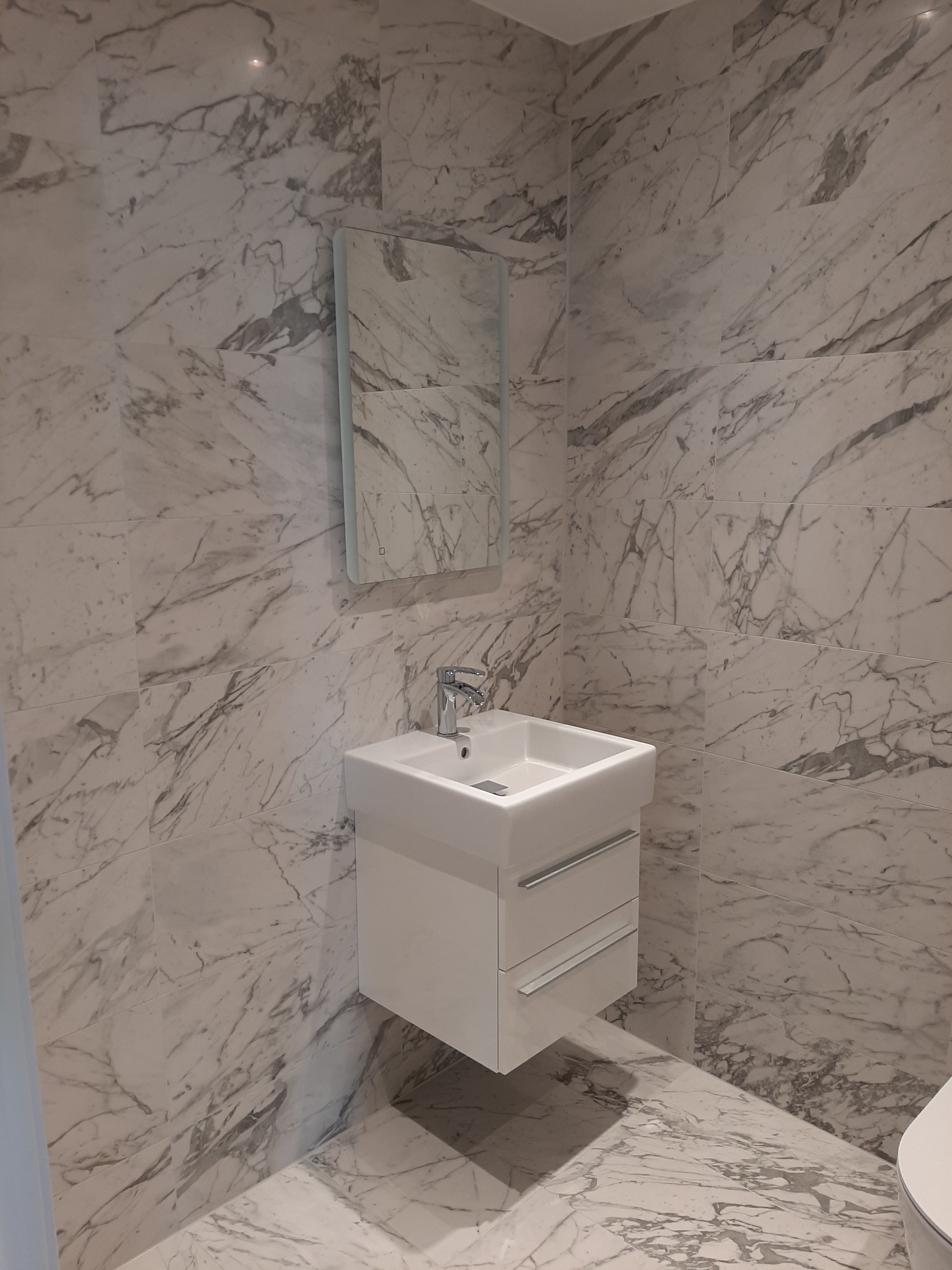 Wykończona łazienka w Brzesku z marmurowymi płytkami na ścianach i podłodze, wisząca szafka z umywalką i lustro z podświetleniem.