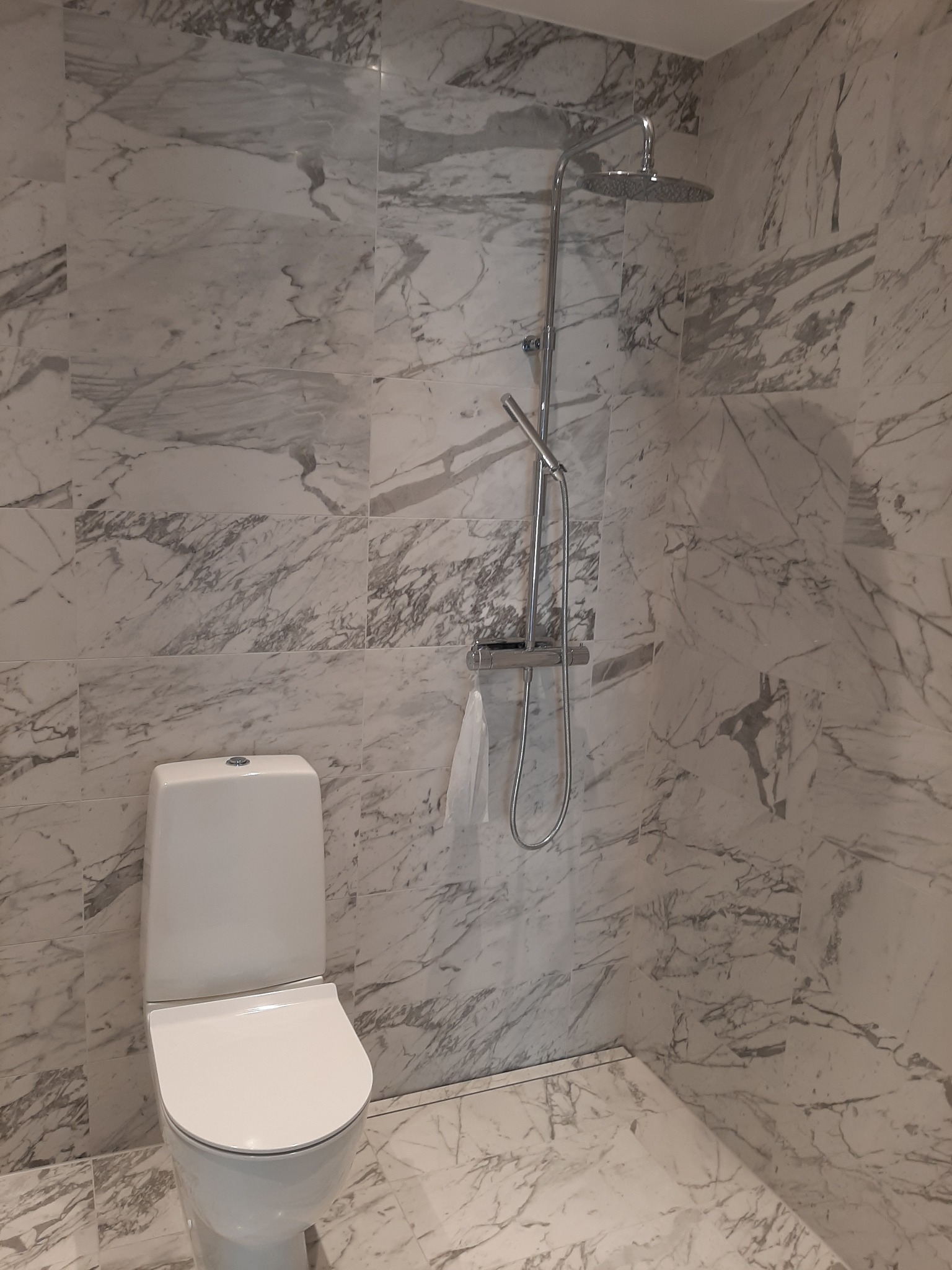 Biała toaleta i chromowana bateria prysznicowa na tle ściany i podłogi z płytek imitujących marmur.