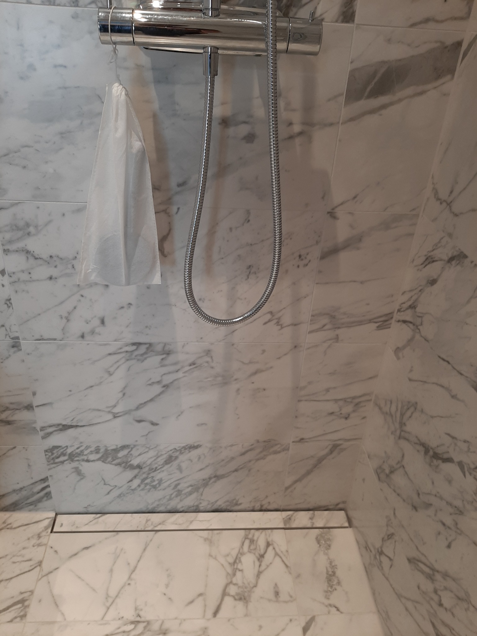 Szczegółowe ujęcie kabiny prysznicowej wyłożonej białym marmurem z szarymi żyłami, widoczny chromowany panel prysznicowy z wężem, mała biała torba zawieszona na panelu oraz odpływ liniowy w podłodze.