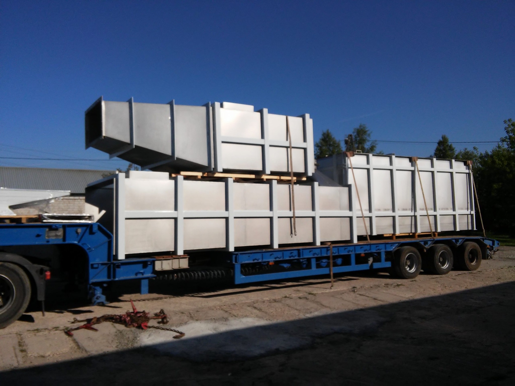 Elementy konstrukcji stalowej, prawdopodobnie moduły wentylacyjne, zabezpieczone na niebieskiej platformie transportowej pod błękitnym niebem.