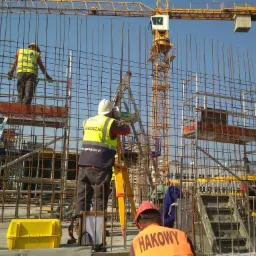 Praca na budowie: Ekipa budowlana w kaskach i kamizelkach odblaskowych, jeden z nich używa niwelatora na żółtym statywie, w tle żelbetowa konstrukcja i żółty dźwig budowlany.