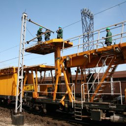 PAK Górnictwo sp. z o.o. - Żółty wagon kolejowy z platformami, na których stoją pracownicy w kaskach, wykonujący prace przy liniach wysokiego napięcia nad torami.