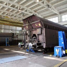 PAK Górnictwo sp. z o.o. - Wnętrze hali z wagonem towarowym na stanowisku naprawczym, pracownik w niebieskim kombinezonie przy wagonie, widoczne podnośniki i skrzynka narzędziowa.