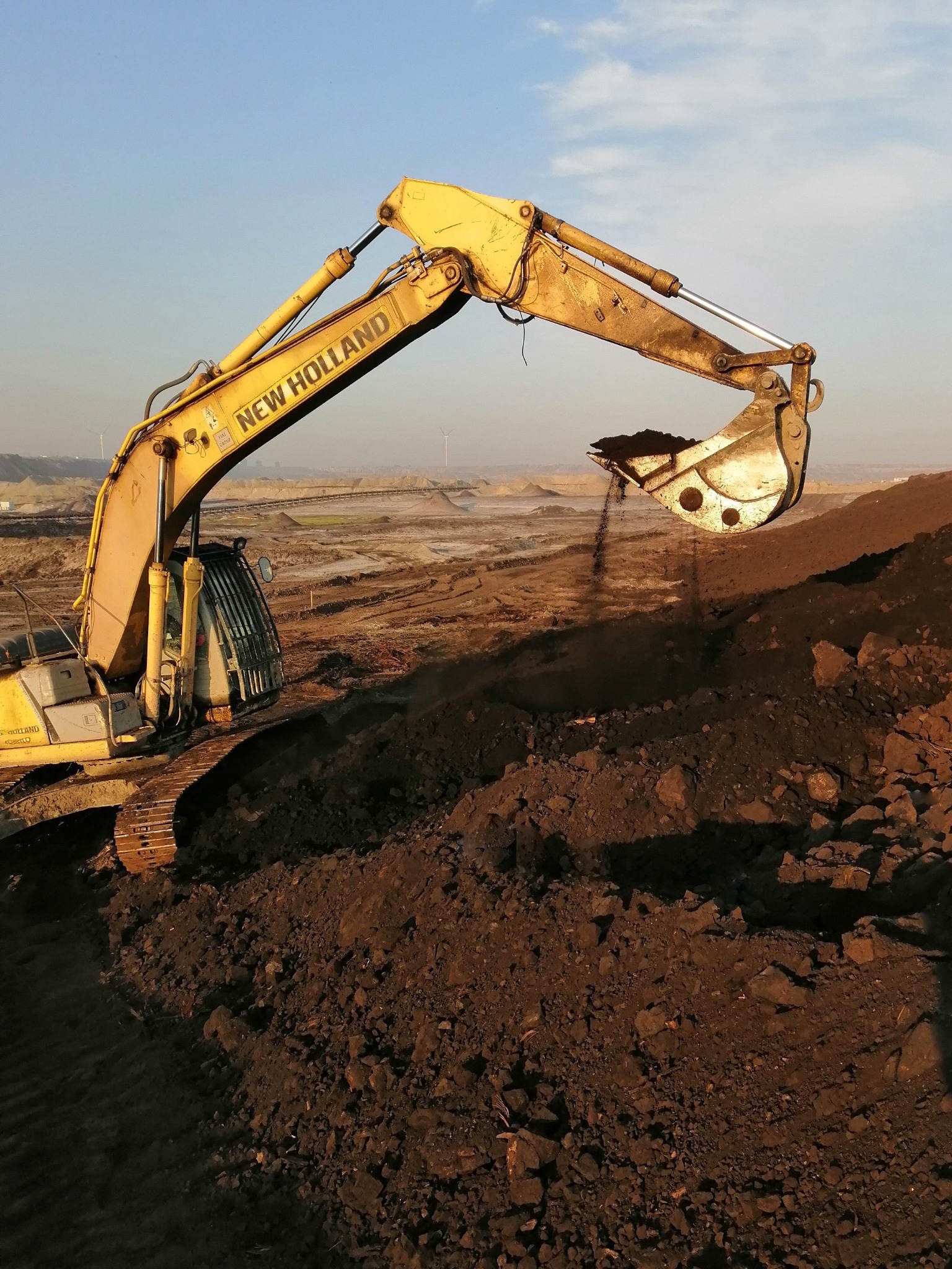 Żółta koparka New Holland nabiera ziemię z wykopu, w tle widoczne wyrównane tereny i turbiny wiatrowe.