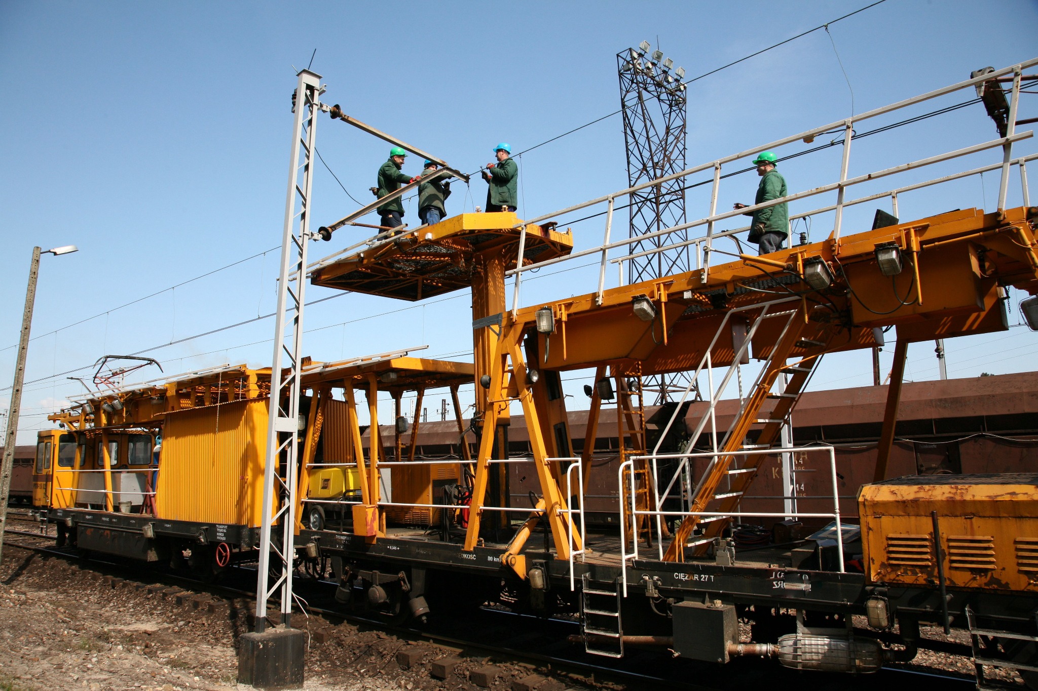 Żółty wagon kolejowy z platformami, na których stoją pracownicy w kaskach, wykonujący prace przy liniach wysokiego napięcia nad torami.