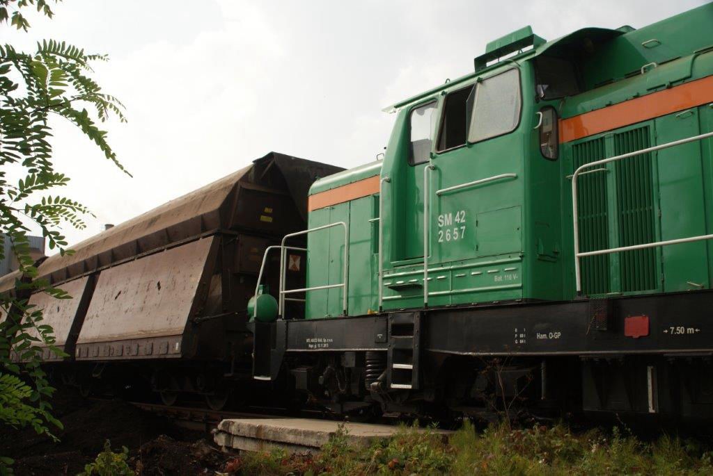 Zielona lokomotywa SM42-2657 z wagonem transportowym wypełnionym ziemią, widoczne detale konstrukcyjne i roślinność w tle.