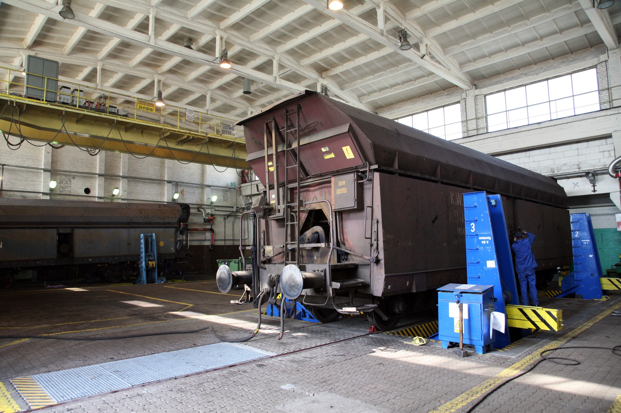 Wnętrze hali z wagonem towarowym na stanowisku naprawczym, pracownik w niebieskim kombinezonie przy wagonie, widoczne podnośniki i skrzynka narzędziowa.
