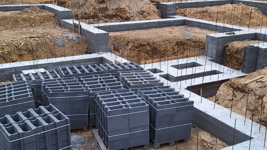 Fundamenty z bloczków betonowych na paletach, widoczne zbrojenia i wykop pod budowę. Ujęcie z góry, ukazujące etap prac fundamentowych.
