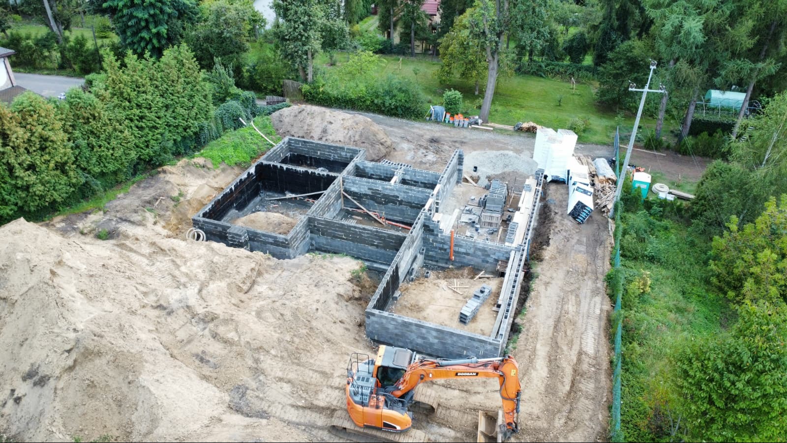 Fundamenty domu jednorodzinnego z izolacją, widok z góry. Na pierwszym planie pomarańczowa koparka. W tle drzewa i zabudowania.