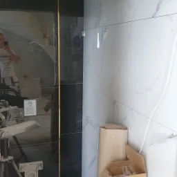 Częściowo wykończona łazienka z czarnymi i białymi płytkami imitującymi marmur, rozdzielonymi złotą listwą. Widoczne narzędzia i materiały budowlane, w tym kartony i zwój kartonu, sugerują trwający...