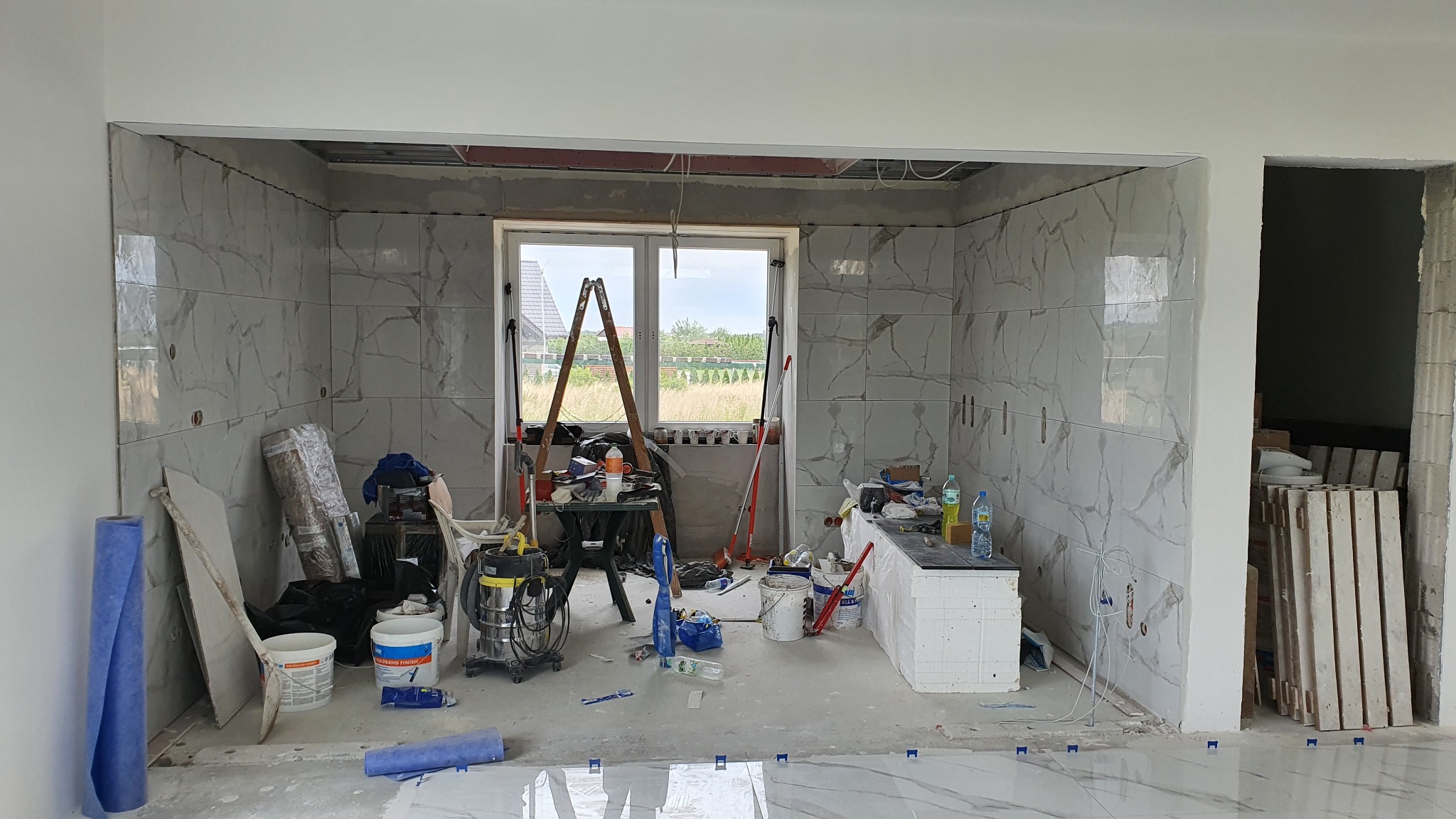 Wnętrze w trakcie układania płytek imitujących marmur na ścianach, widoczne narzędzia glazurnicze, drabina oparta o okno i częściowo ułożona podłoga z płytek.