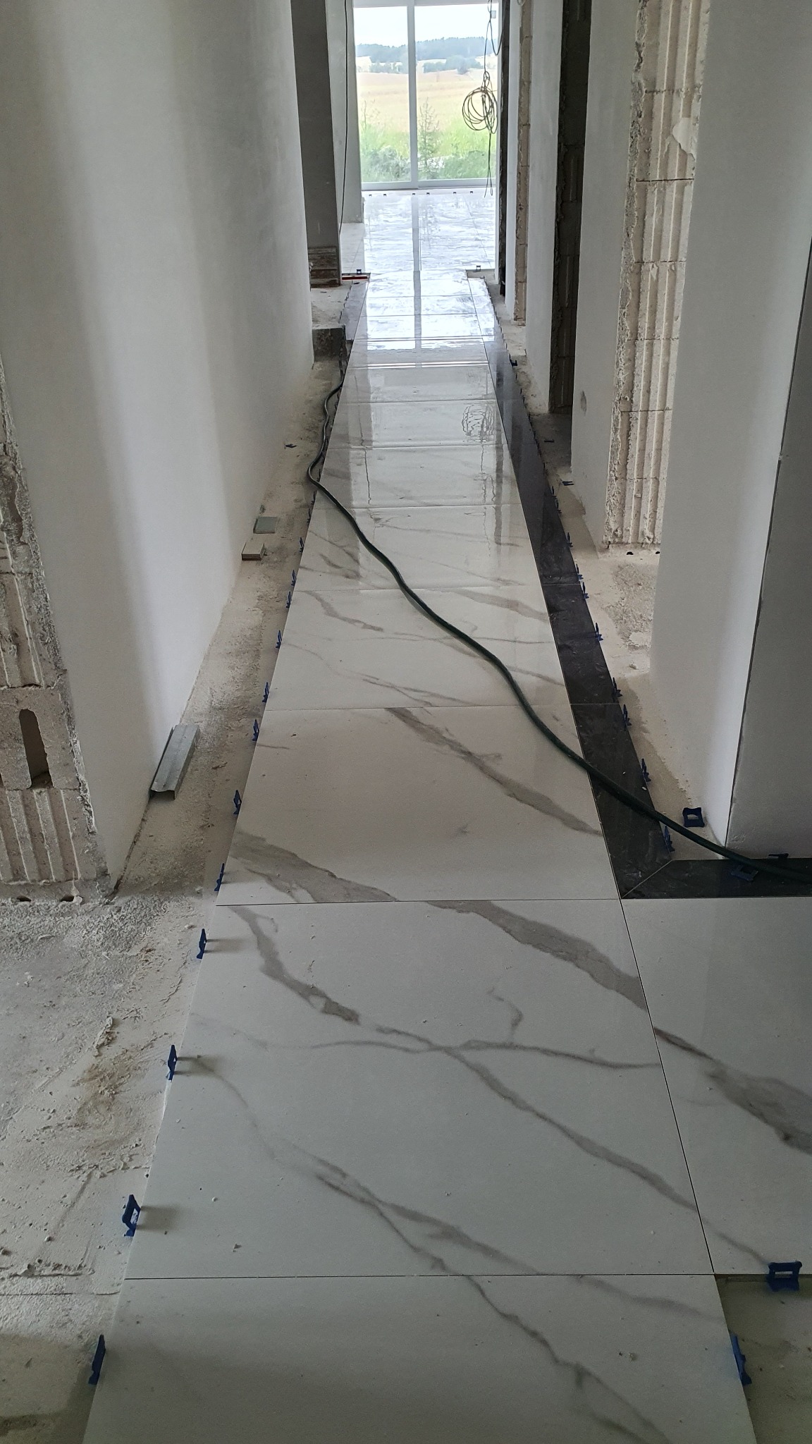 Korytarz w trakcie układania białych płytek imitujących marmur, z widocznymi niebieskimi klipsami poziomującymi i czarnym kablem na powierzchni.