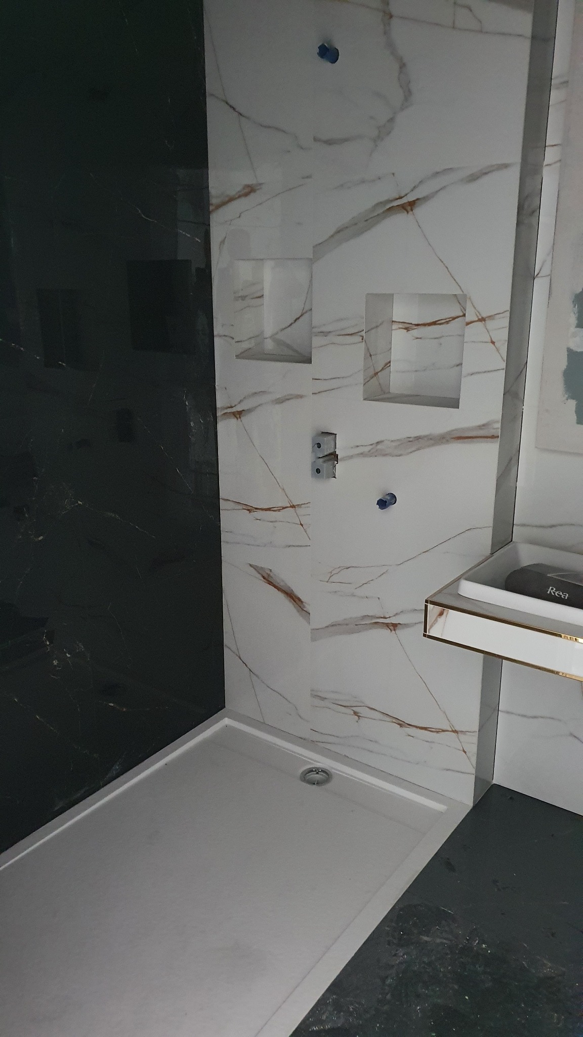 Nowoczesna kabina prysznicowa z czarną ścianą i białymi płytkami imitującymi marmur z złotymi żyłkami; widoczne wnęki w ścianie i brodzik z odpływem liniowym.