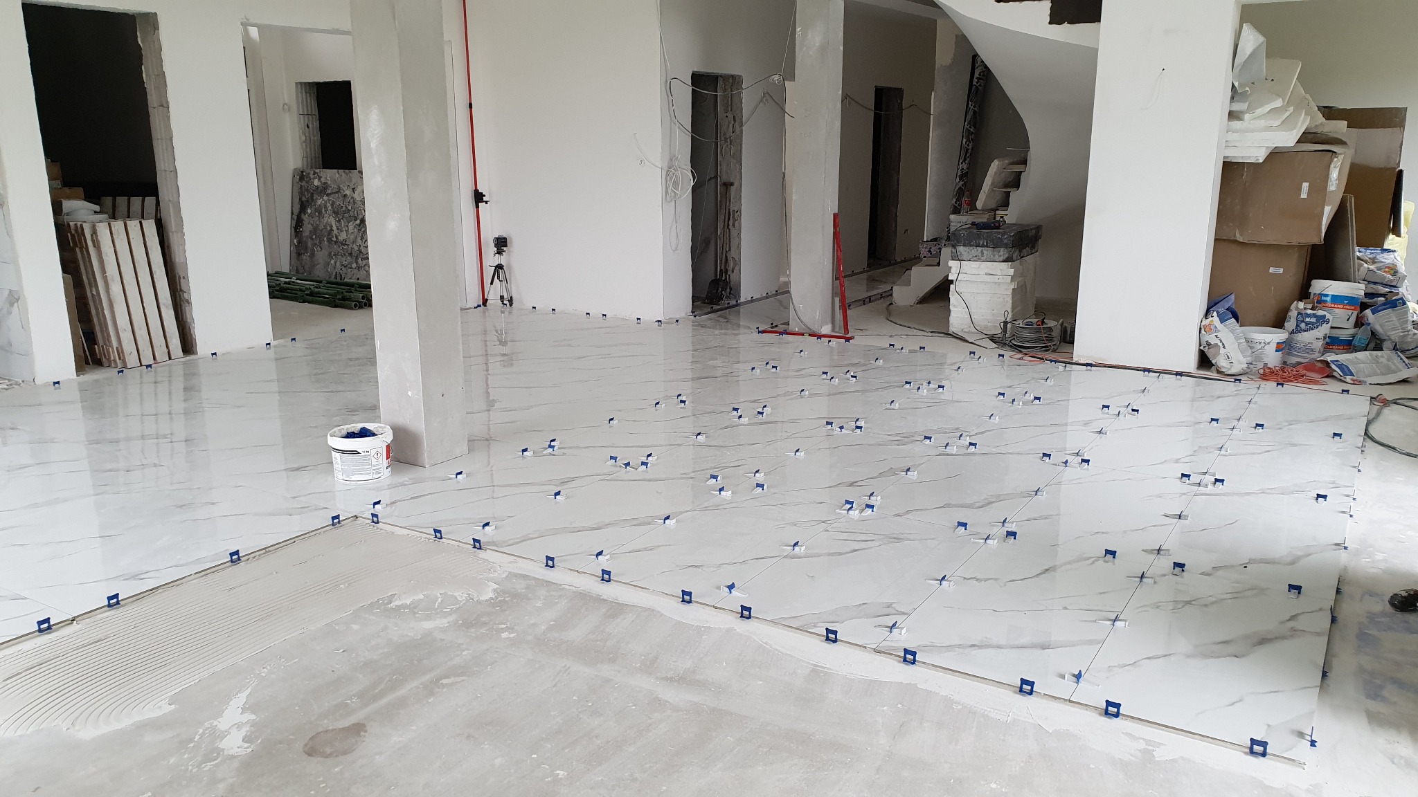 Układanie białych płytek podłogowych z marmurkowym wzorem, z użyciem niebieskich klipsów poziomujących, w surowym wnętrzu z widocznymi instalacjami i materiałami budowlanymi.