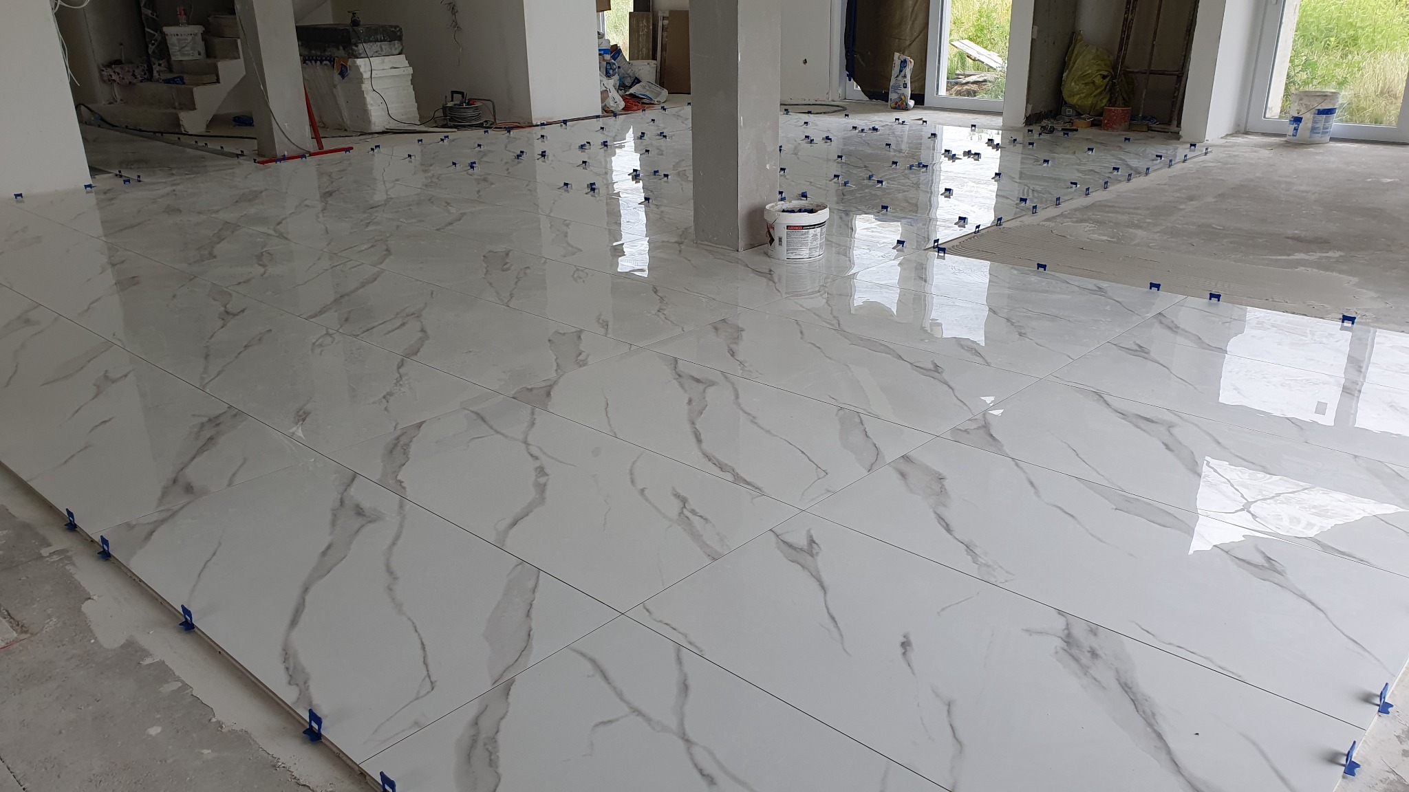 Układanie białych płytek podłogowych z szarym marmurkowym wzorem, użycie klipsów poziomujących, widoczny etap prac wykończeniowych.