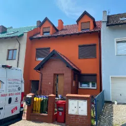 Pomarańczowy dom jednorodzinny z ceglanym gankiem i zamkniętymi roletami zewnętrznymi, widziany z poziomu chodnika w słoneczny dzień.