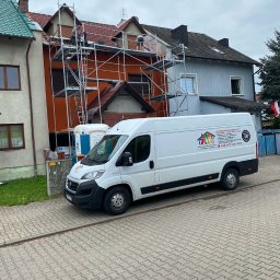Usługi Budowlane Andrzej Świerc - Pracownicy na rusztowaniu podczas renowacji elewacji budynku z pomarańczową fasadą. Na pierwszym planie biały samochód dostawczy z logo firmy.
