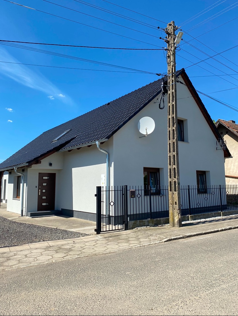 Jasny, jednopiętrowy dom z ciemnym dachem, okna z brązowymi ramami, biała satelitarna antena na ścianie, czarny metalowy płot i betonowy słup energetyczny na pierwszym planie, błękitne niebo w tle.