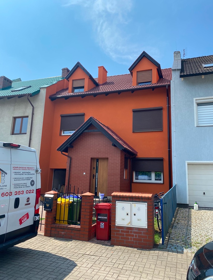 Pomarańczowy dom jednorodzinny z ceglanym gankiem i zamkniętymi roletami zewnętrznymi, widziany z poziomu chodnika w słoneczny dzień.