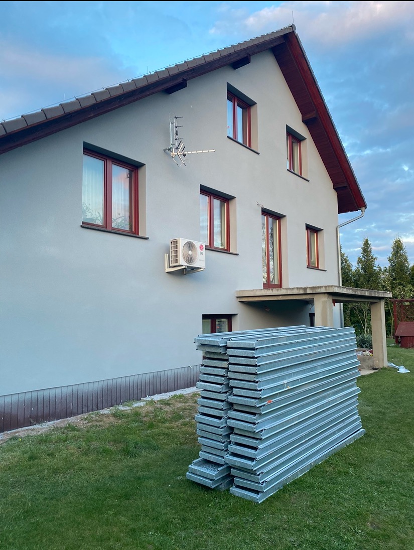 Dwupiętrowy dom z szarą elewacją i brązowymi ramami okiennymi, na trawniku stos metalowych paneli przed montażem, widoczny klimatyzator LG na ścianie i antena satelitarna.