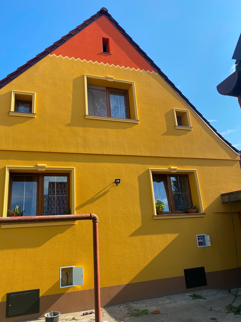 Żółta elewacja domu jednorodzinnego z pomarańczowym szczytem i dekoracyjnym zygzakiem, widoczne okna z roślinami na parapetach, rura gazowa przy ścianie, skrzynka elektryczna oraz wiadro na ziemi.