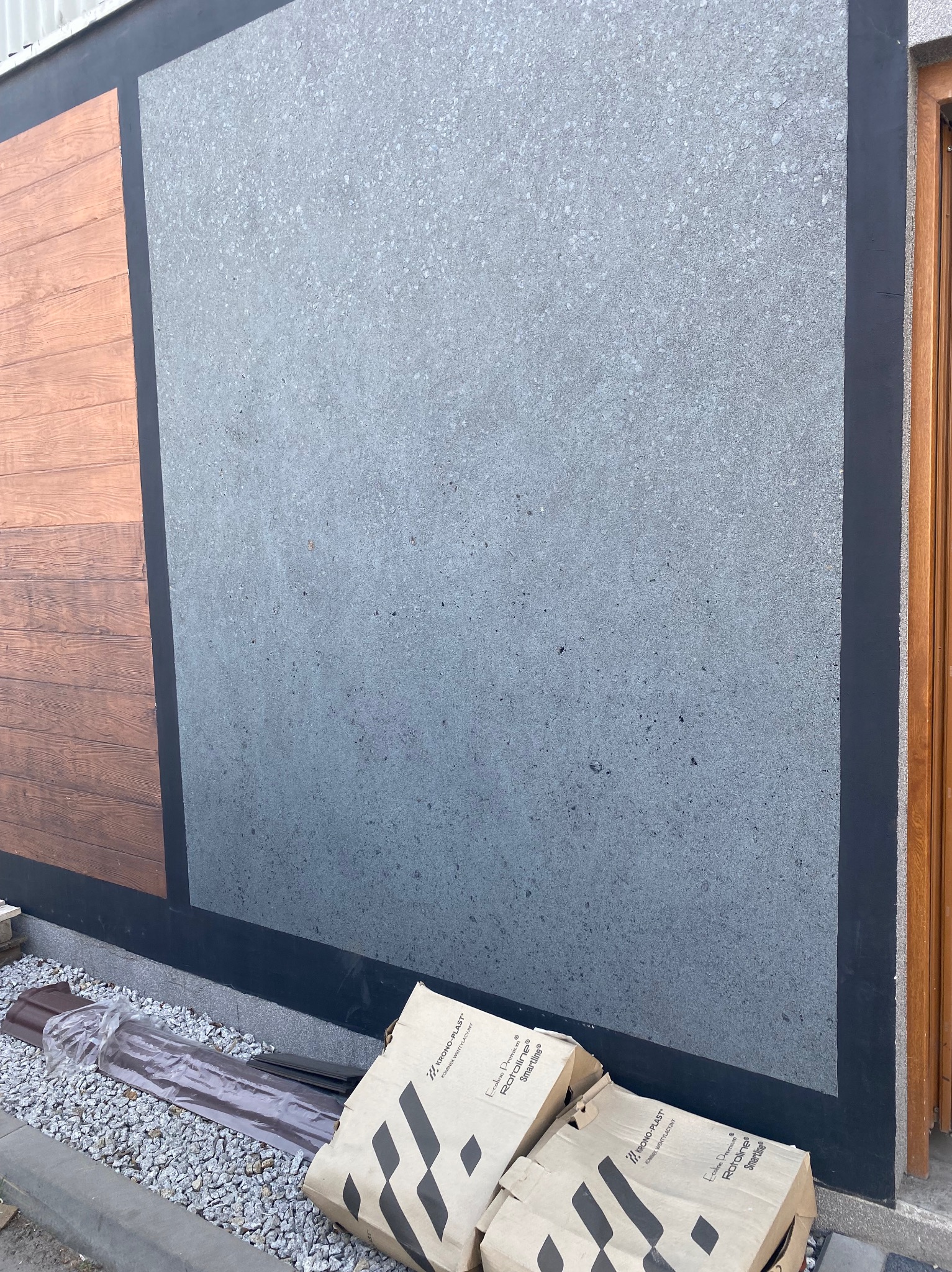 Fragment elewacji z szarą fakturą, obok drewnopodobna okładzina. U dołu worki z zaprawą i elementy wentylacyjne. Czarny pas oddziela faktury.