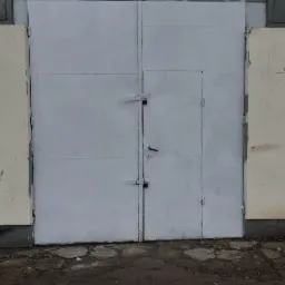 Szare, metalowe wrota warsztatu samochodowego z szyldem 'BK AUTO' i numerem telefonu, widok z zewnątrz na nierówny chodnik z sześciokątnych płyt.