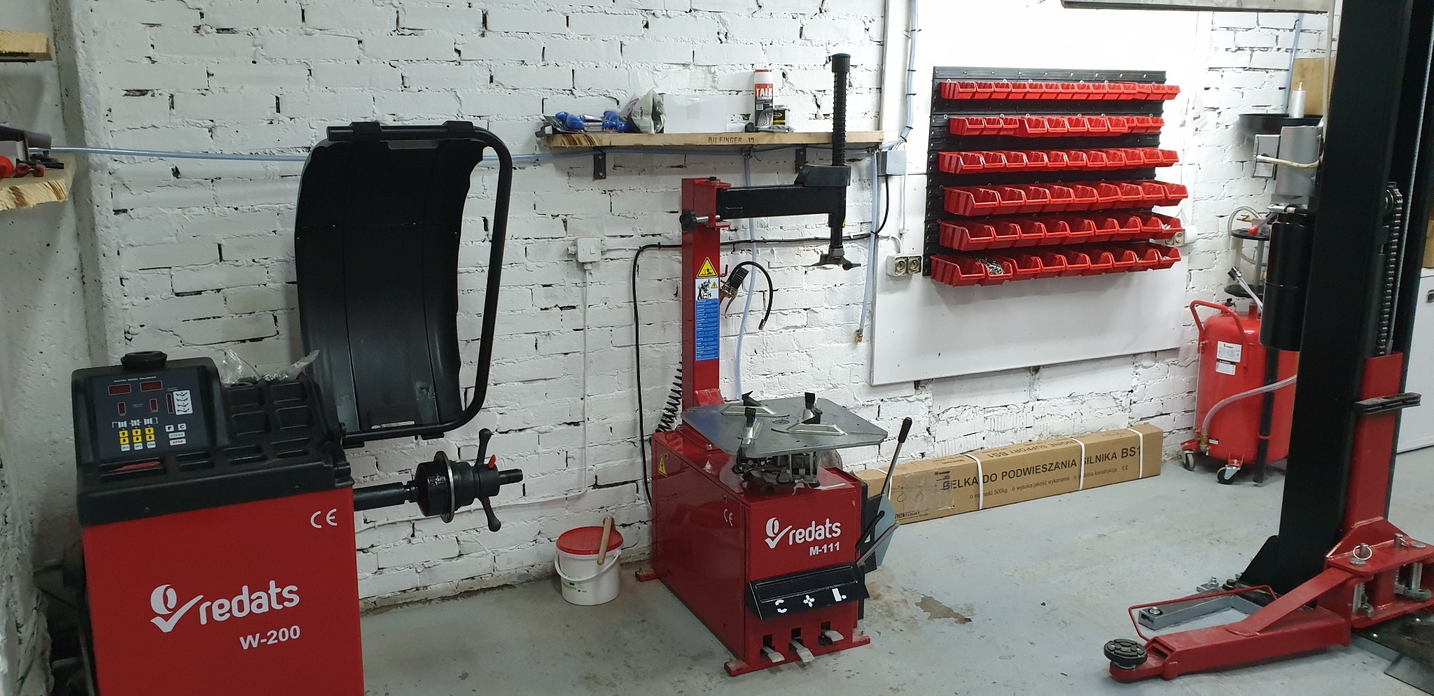 Wyposażenie warsztatu w postaci wyważarki do kół Redats W-200, montażownicy do opon Redats M-111 oraz podnośnika dwukolumnowego, na tle białej ściany z cegły i półki z narzędziami.