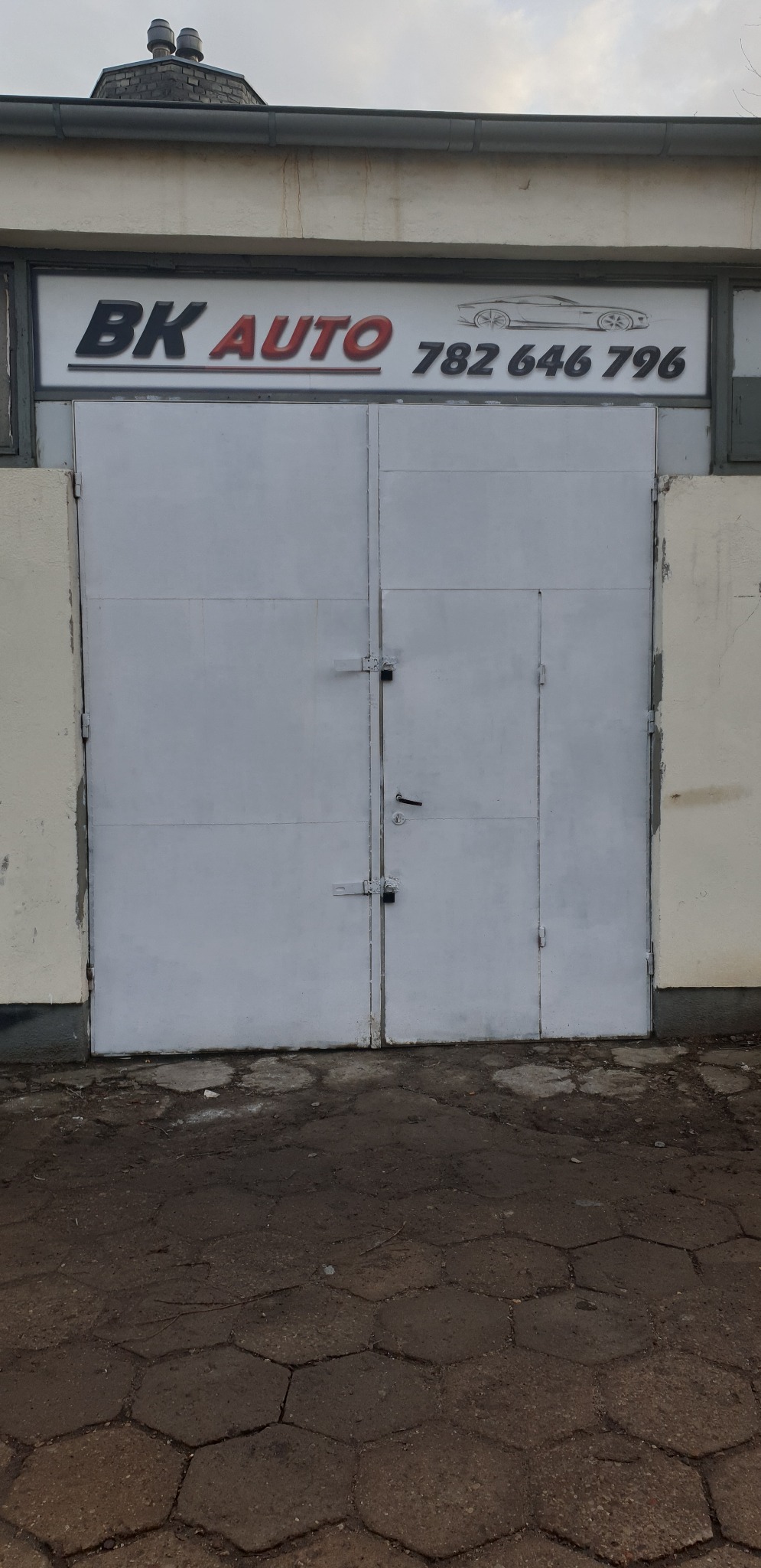 Szare, metalowe wrota warsztatu samochodowego z szyldem 'BK AUTO' i numerem telefonu, widok z zewnątrz na nierówny chodnik z sześciokątnych płyt.