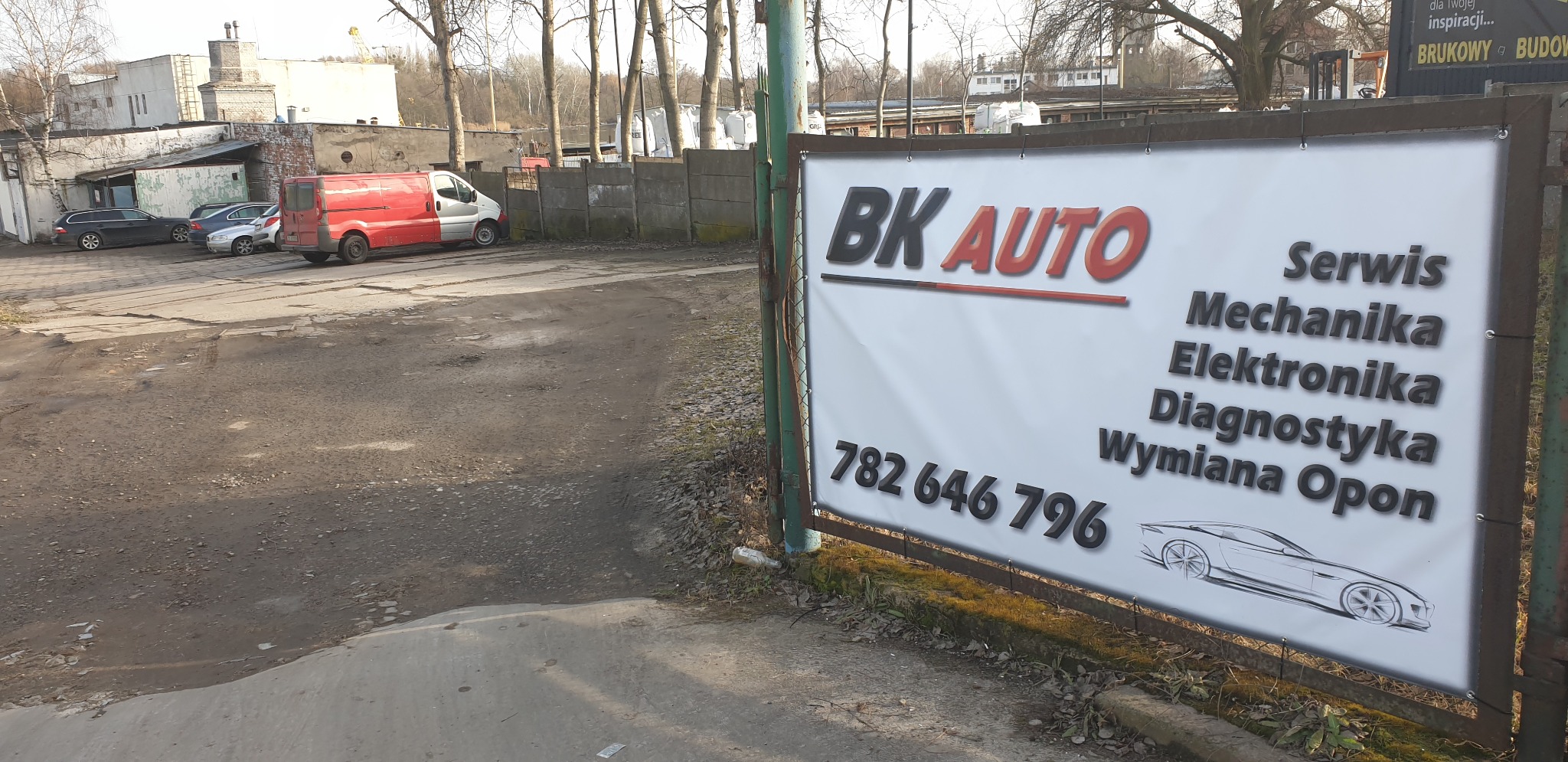 Tablica reklamowa warsztatu 'BK AUTO' z ofertą usług: mechanika, elektronika, diagnostyka, wymiana opon, widoczna na tle placu z zaparkowanymi samochodami i budynkami.