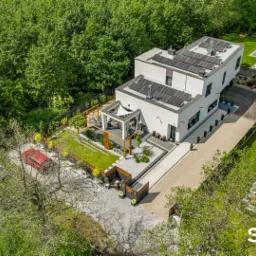 Nowoczesny dom z białymi ścianami i panelami słonecznymi na dachu, widok z lotu ptaka na zadbany ogród z pergolą i dwoma samochodami zaparkowanymi na posesji.