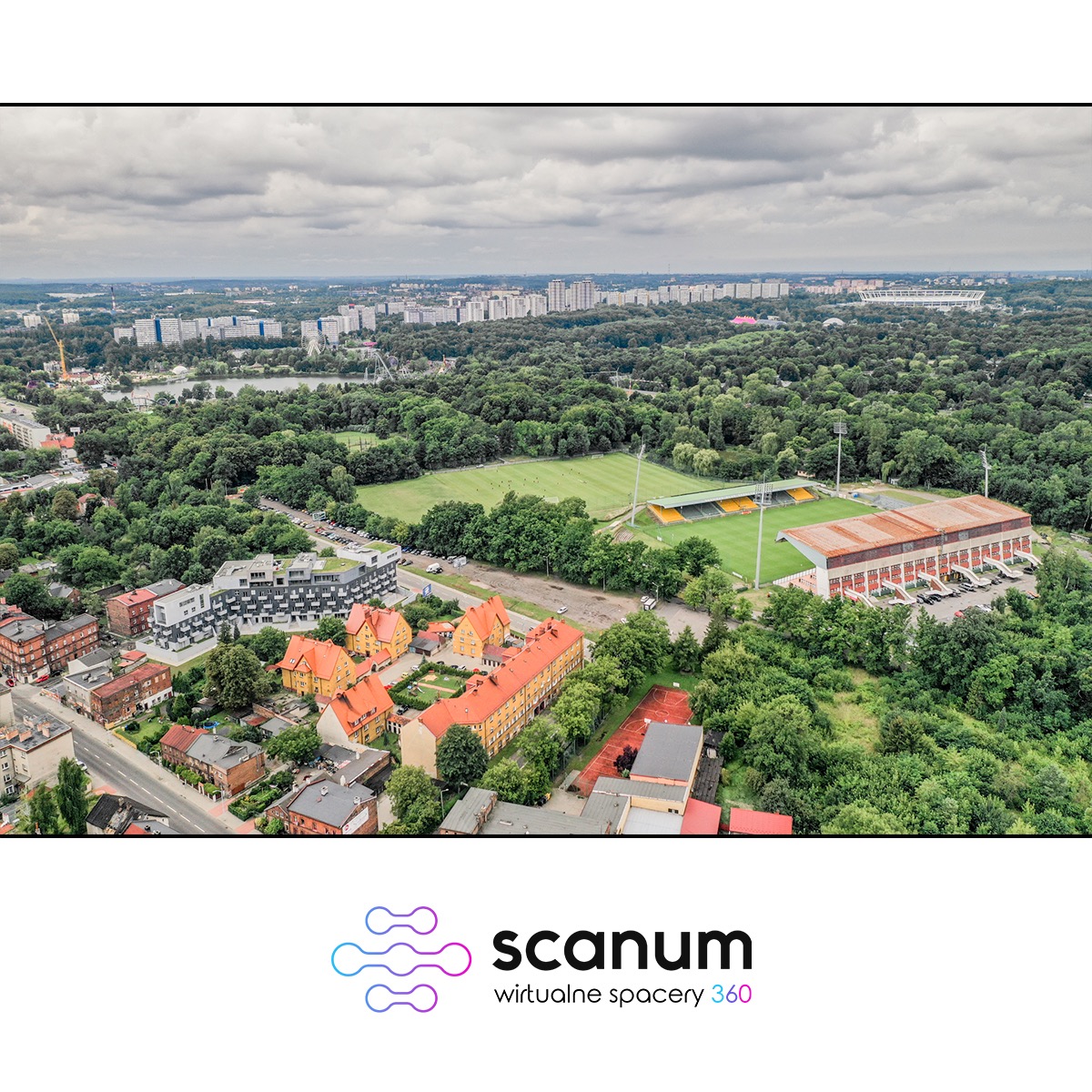 Panorama miasta z lotu ptaka, widoczne budynki mieszkalne, stadion piłkarski otoczony zielenią, pochmurne niebo.