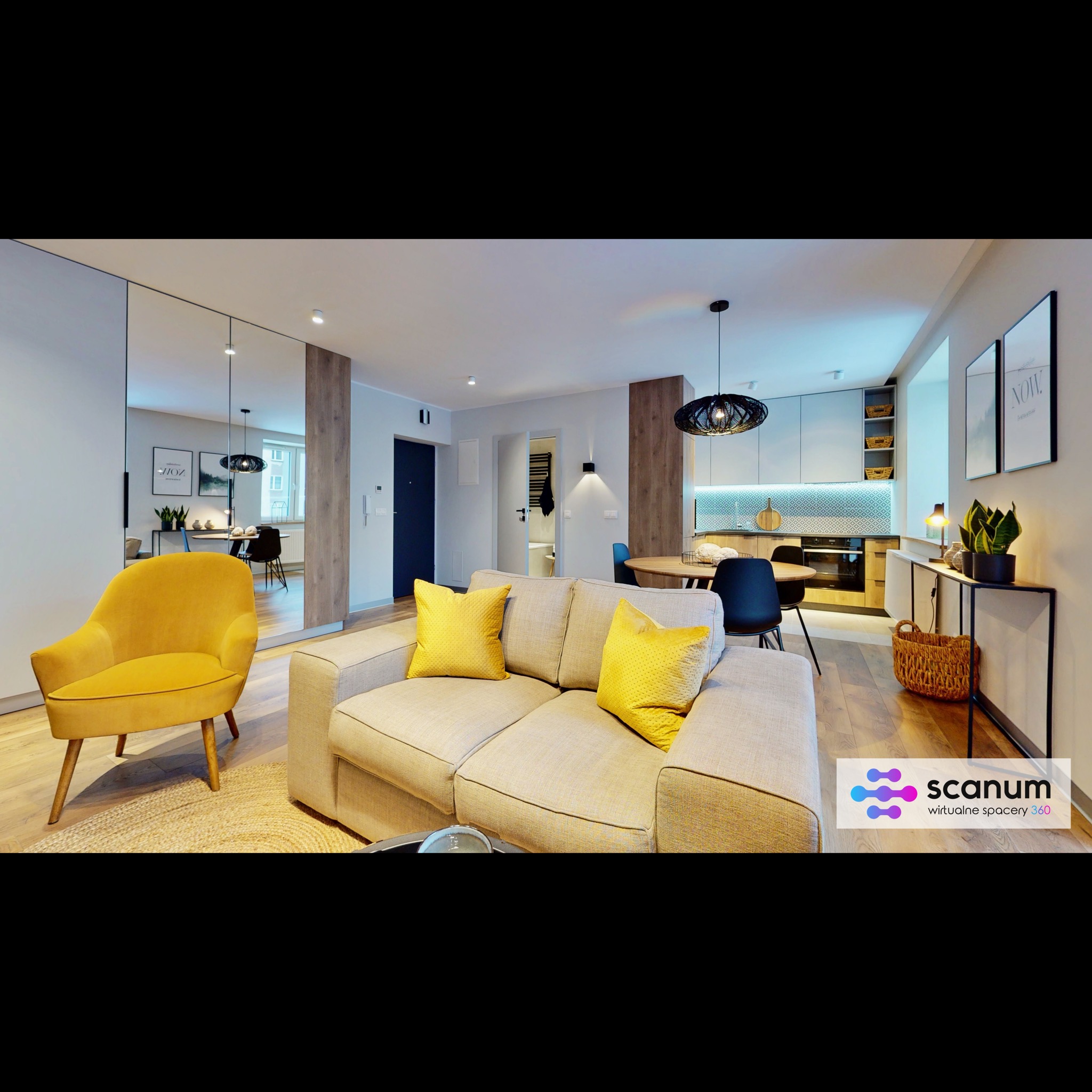 Nowoczesne wnętrze mieszkania z żółtym fotelem i kanapą, widok na kuchnię z okrągłym stołem, lustro na ścianie powiększające przestrzeń, drewniana podłoga i logo firmy Scanum.