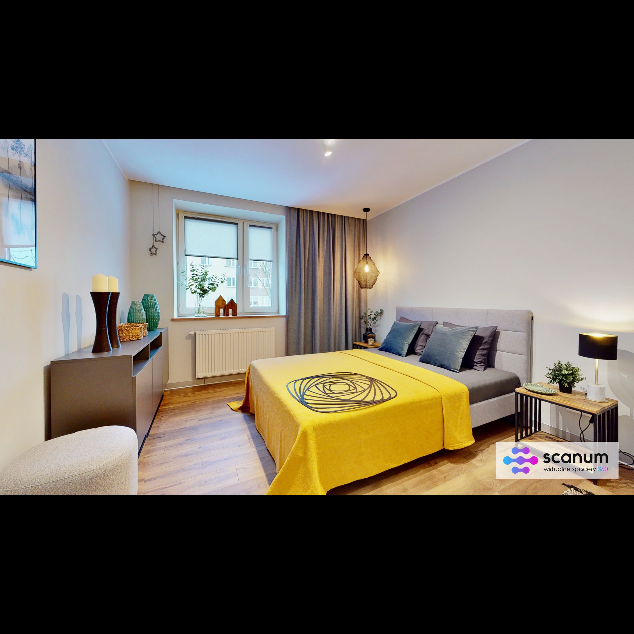 Jasna sypialnia z drewnianą podłogą, szarą komodą, łóżkiem z szarą ramą i żółtą narzutą z geometrycznym wzorem, oświetlona lampą nocną i wiszącą lampą, z widokiem na okno z roletami.