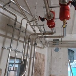 Usługi instalacyjne Michał Smektalski - Skomplikowany układ rur ze stali nierdzewnej, z czerwonymi elementami, zamontowany pod sufitem, w surowym wnętrzu budynku w trakcie instalacji.