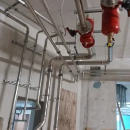 Skomplikowany układ rur ze stali nierdzewnej, z czerwonymi elementami, zamontowany pod sufitem, w surowym wnętrzu budynku w trakcie instalacji.