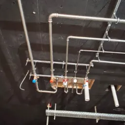 Kompleksowa instalacja hydrauliczna i grzewcza z rur miedzianych, zamontowana pod sufitem, widoczne liczne zawory i przyłącza. Czarny sufit i elementy konstrukcyjne.