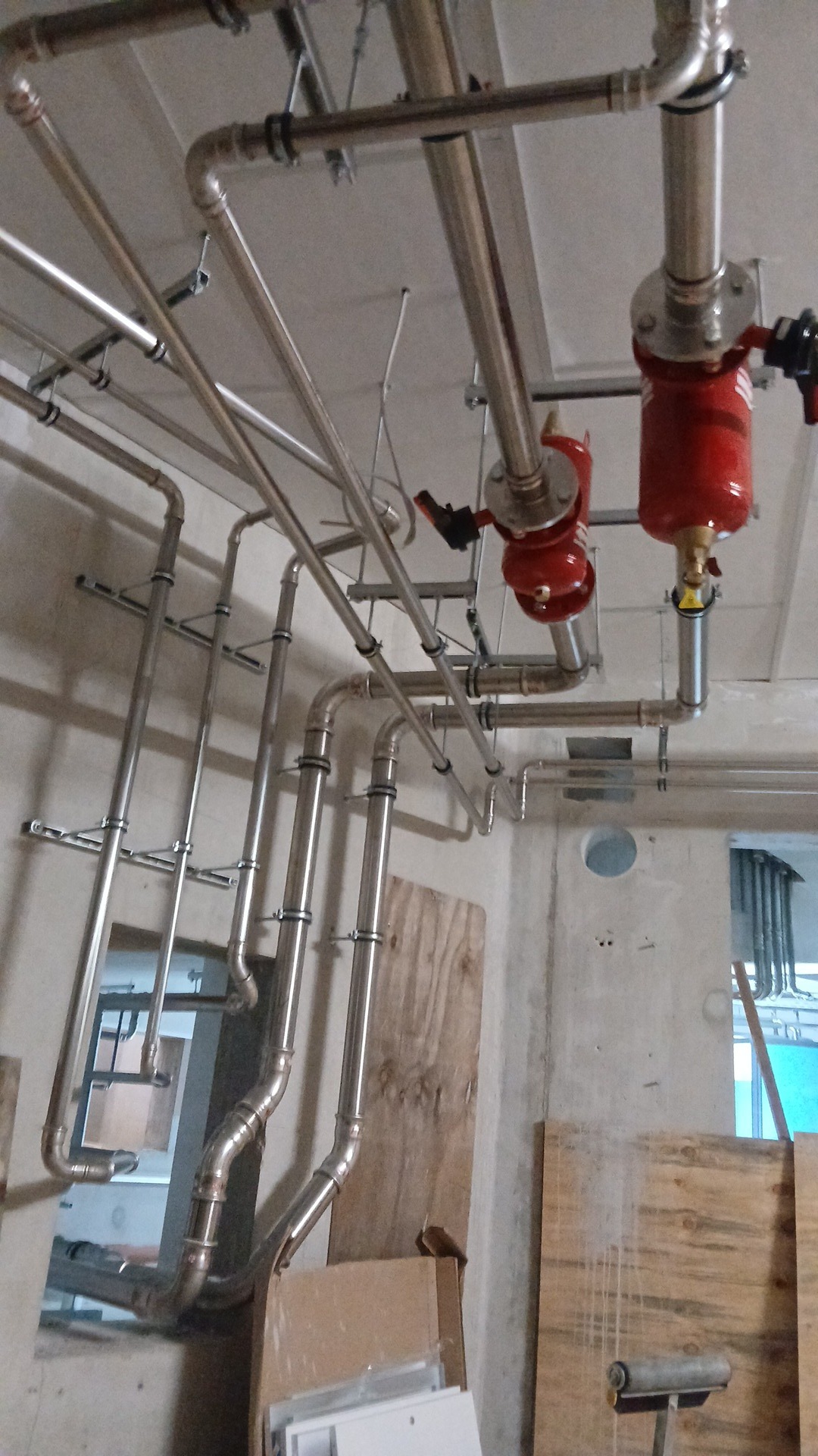 Skomplikowany układ rur ze stali nierdzewnej, z czerwonymi elementami, zamontowany pod sufitem, w surowym wnętrzu budynku w trakcie instalacji.