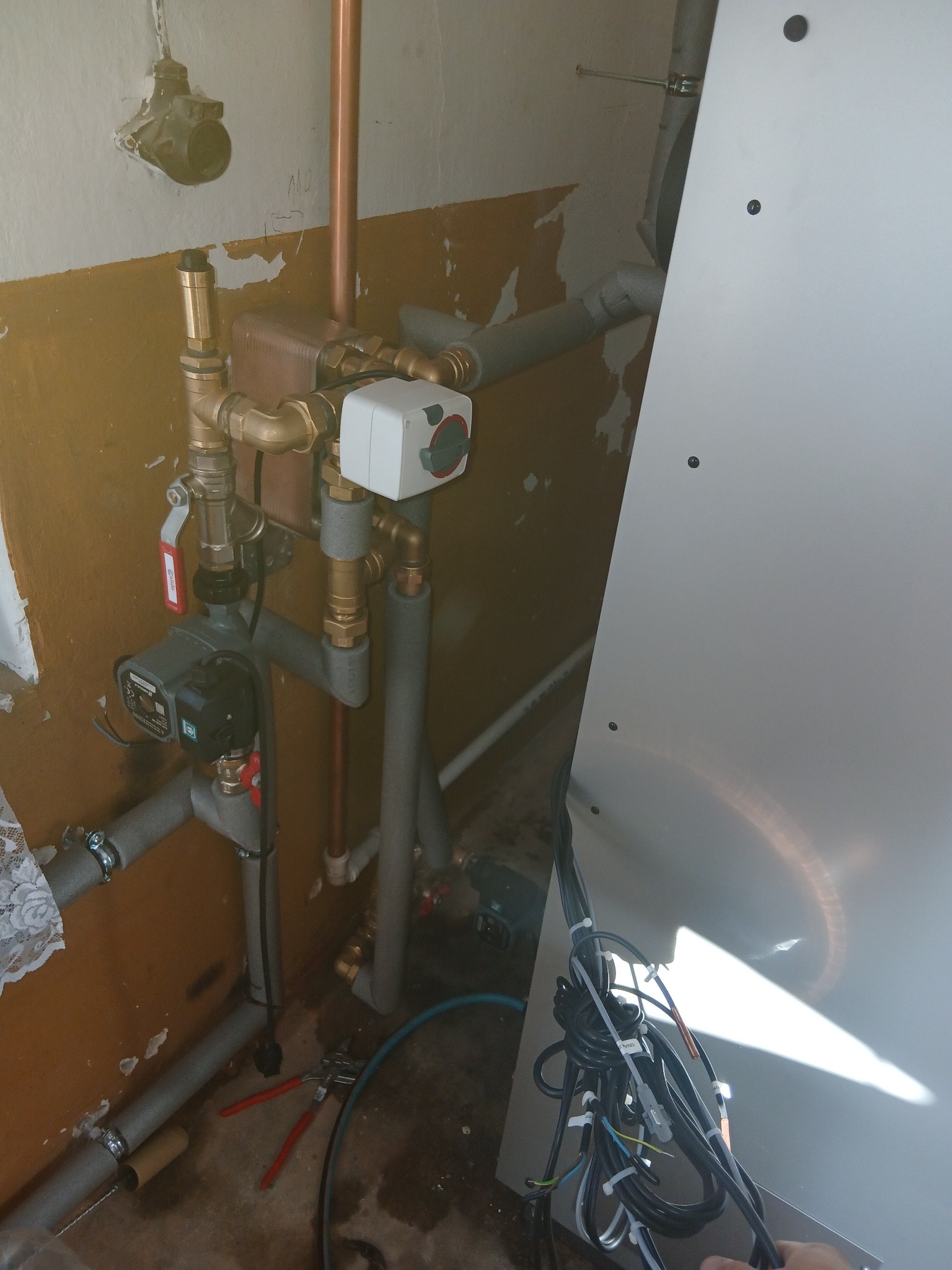 Węzeł cieplny z pompą obiegową i zaworami, rury miedziane i szare izolowane, w tle fragment ściany z instalacją elektryczną. Widoczne narzędzia na podłodze.