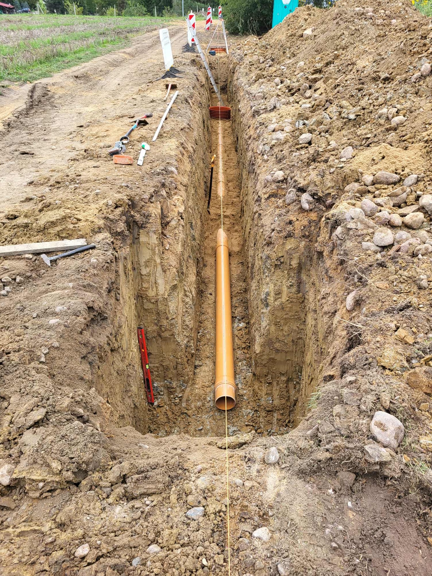 Pomarańczowa rura kanalizacyjna w wykopie ziemnym, poziomica, narzędzia i zabezpieczenia wykopu widoczne na powierzchni. Prace ziemne w trakcie realizacji.