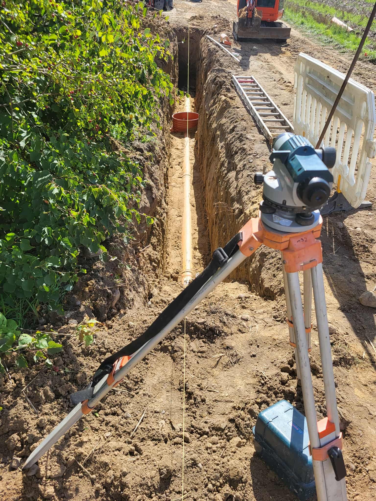 Wykop z ułożonym rurociągiem, widoczny niwelator optyczny na statywie, drabina i bariera. W tle koparka i zieleń. Prace ziemne przyłącza wod-kan w słoneczny dzień.