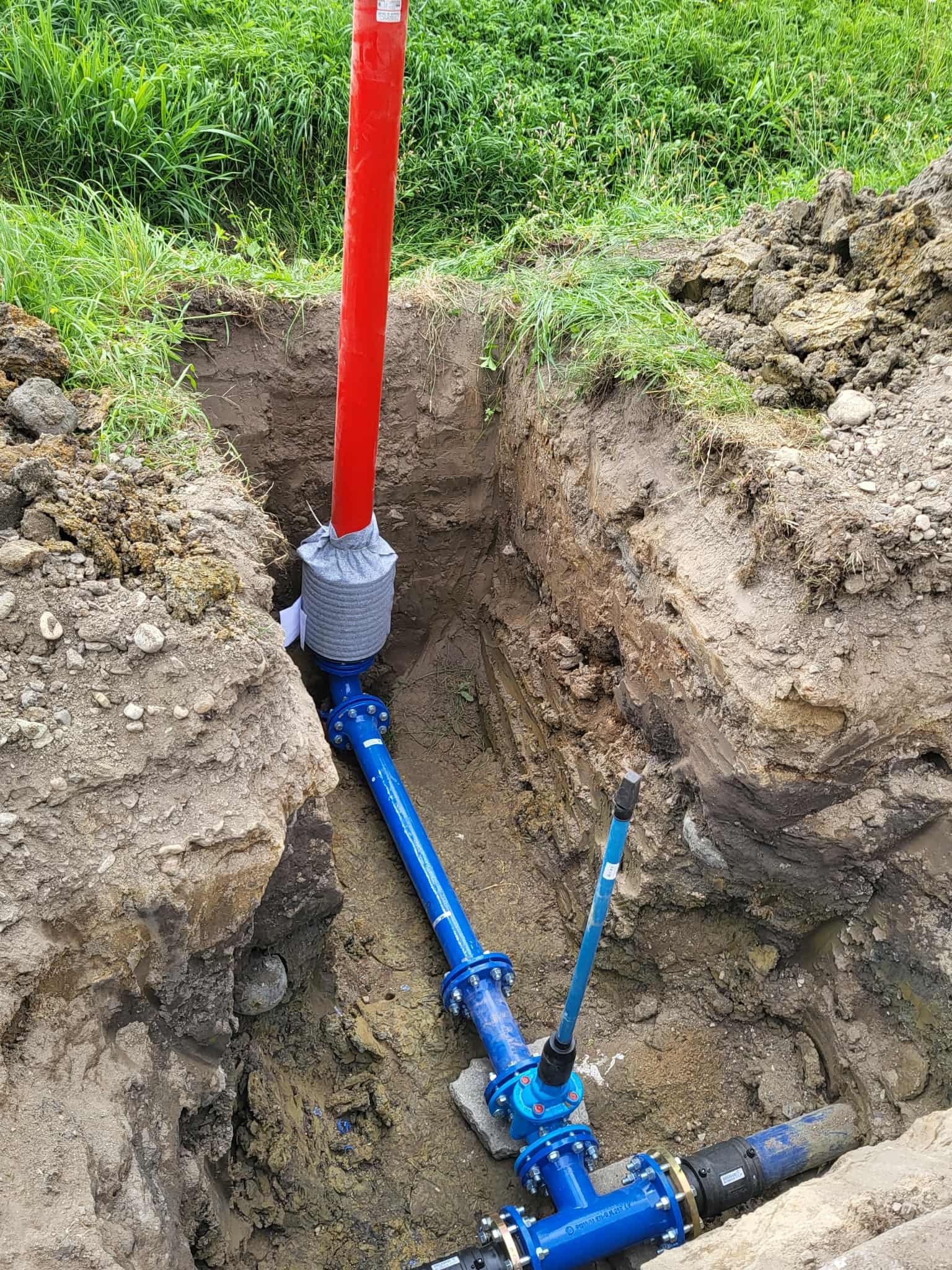 Niebieska instalacja wodna w wykopie ziemnym z czerwoną rurą pionową. Elementy złączone śrubami, widoczne narzędzia i otoczenie z trawą.