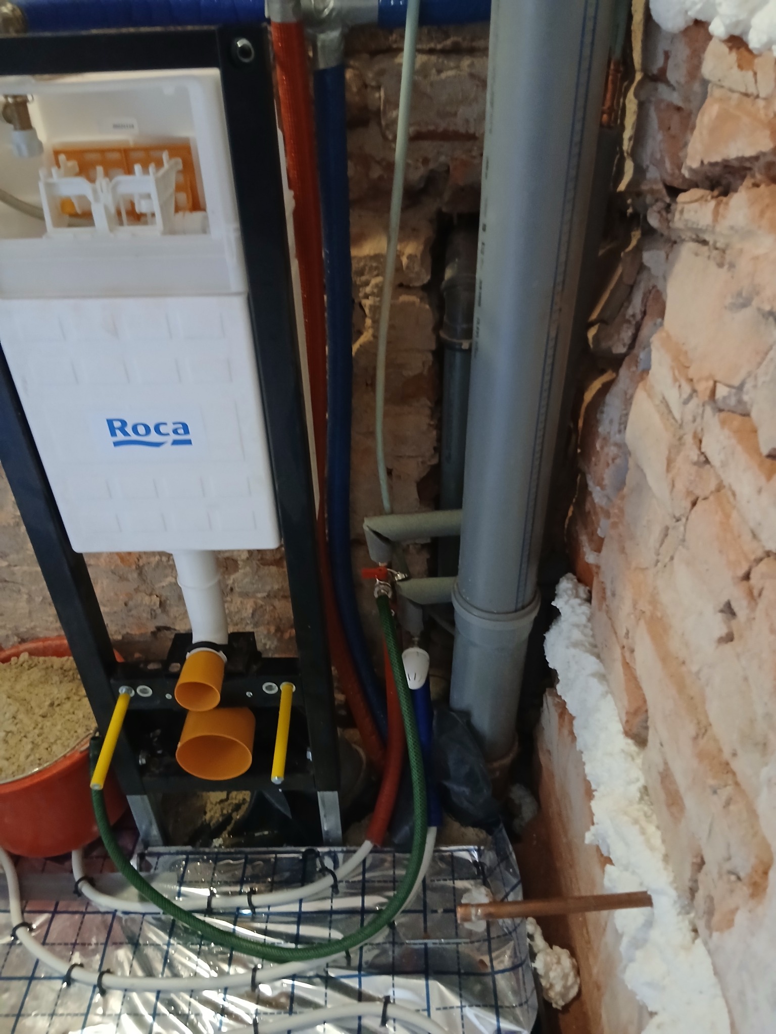 Montaż podtynkowej spłuczki Roca, instalacja ogrzewania podłogowego, rury wodne w surowym wnętrzu z ceglaną ścianą. Widoczne materiały i narzędzia.