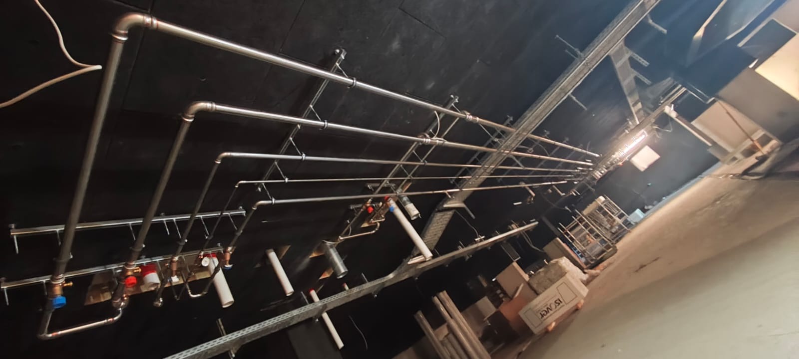 Instalacja rur miedzianych pod sufitem w ciemnym pomieszczeniu, widoczne elementy mocujące i rozgałęzienia, instalacja częściowo ukończona, widoczne materiały budowlane.