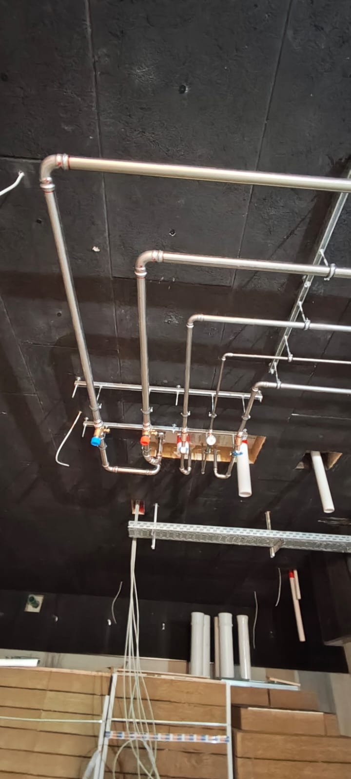 Kompleksowa instalacja hydrauliczna i grzewcza z rur miedzianych, zamontowana pod sufitem, widoczne liczne zawory i przyłącza. Czarny sufit i elementy konstrukcyjne.
