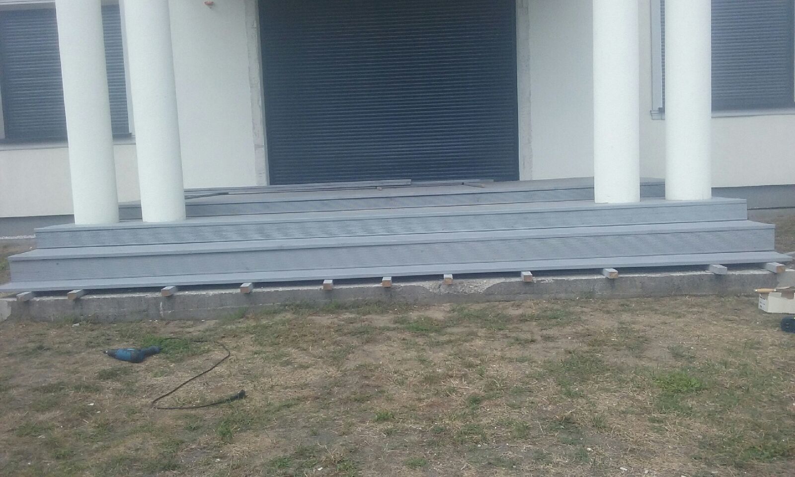 Montaż szarych desek kompozytowych na schodach wejściowych do budynku z białymi kolumnami, widoczne narzędzia i elementy konstrukcyjne w trakcie prac.