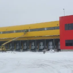 Pracownik na podnośniku koszowym czyści okna na wysokości żółtego budynku magazynowego z licznymi bramami załadunkowymi; widoczny czerwony fragment budynku, zimowa sceneria.