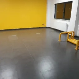 Puste biuro z szarą podłogą, żółtą ścianą, białymi ścianami, grzejnikami i żółtymi barierkami ochronnymi.